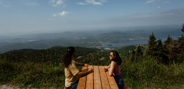 a couple smiling at each other on top of the Mont Tremblant, Quebec mountain after a stunning hike during a summer getaway